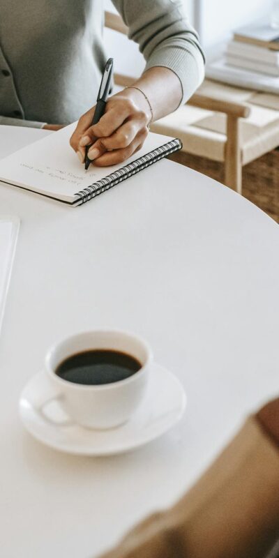 From above crop multiracial female interviewer or psychologist in formal wear asking questions and taking notes in planner while talking to black man and sitting together at table with coffee