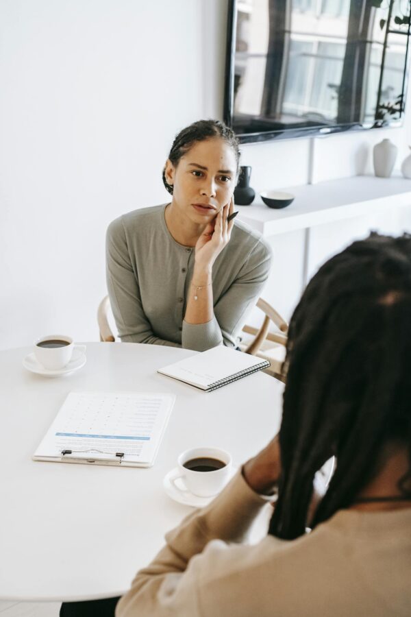 Serious ethnic female consultant listening to black male client problems while sitting at round table together in light psychology center